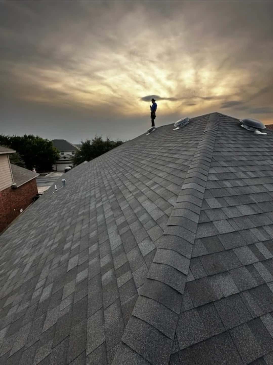 Silhouette of a roofer standing on a finished gray shingle ridge at sunset.