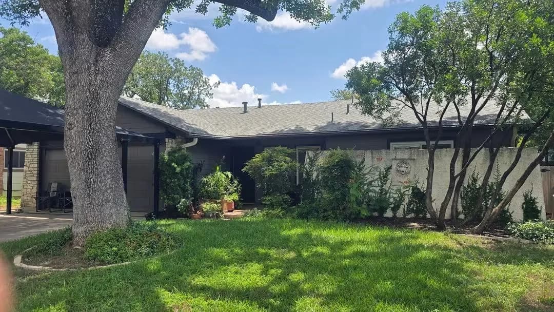 Single-story dark stone home with a completed gray shingle roof shaded by a large oak tree.