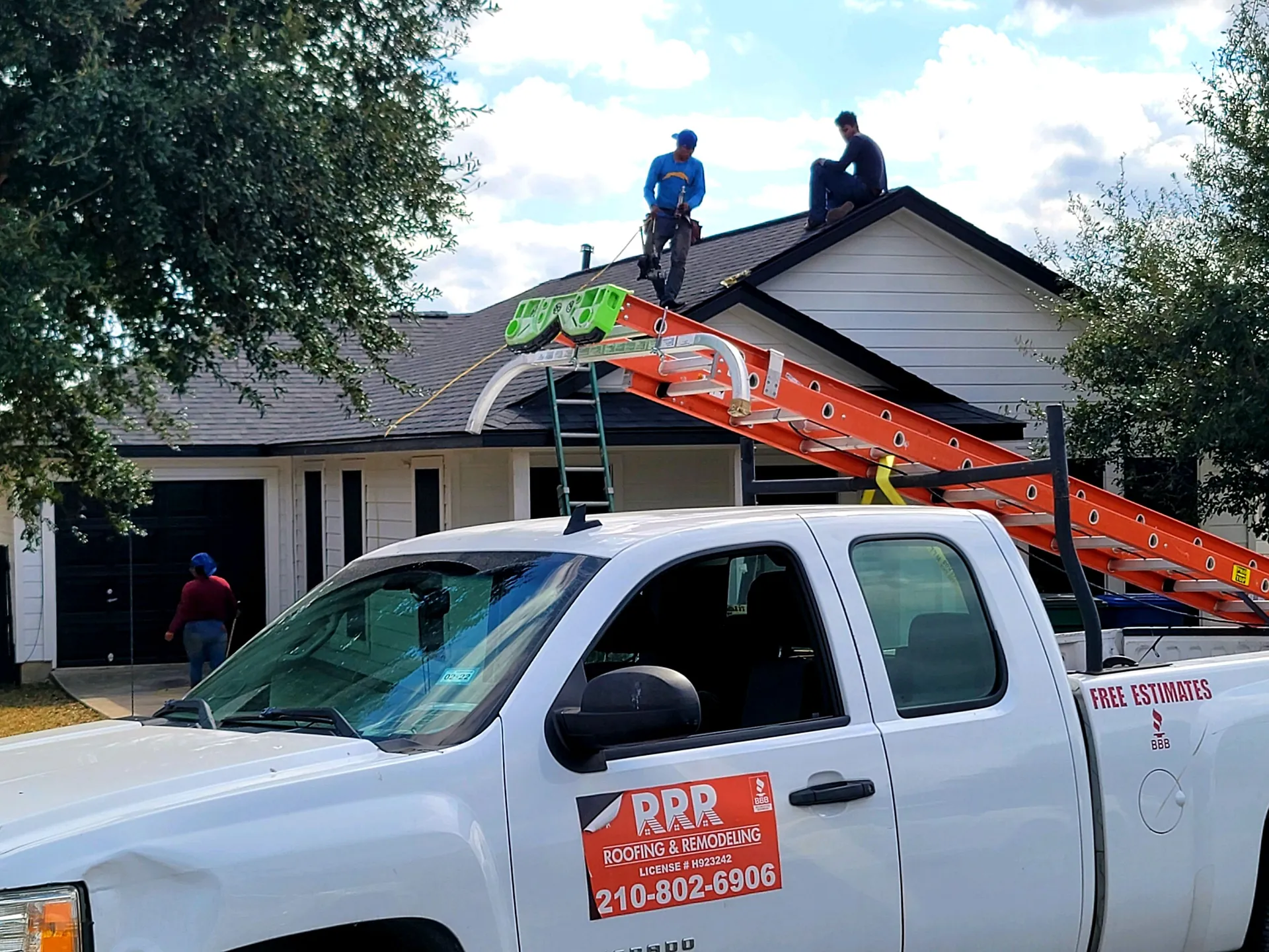 White company pickup with an RRR door magnet and ladder rack parked at a home with two roofers on the roof.